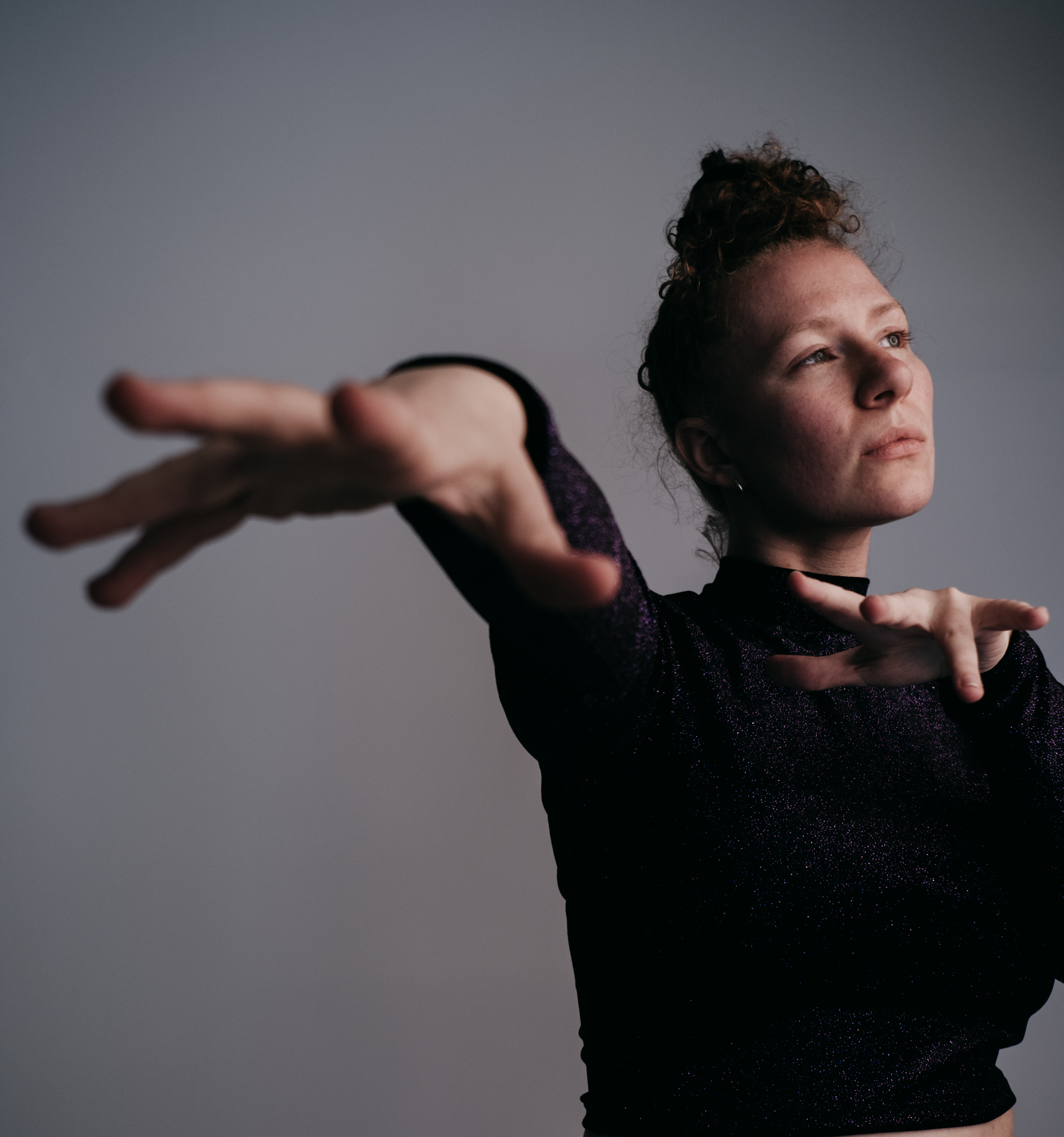 Jemma Freese stood in front of a grey background looking off to the right. Her hands and fingers are outstretched towards the camera.