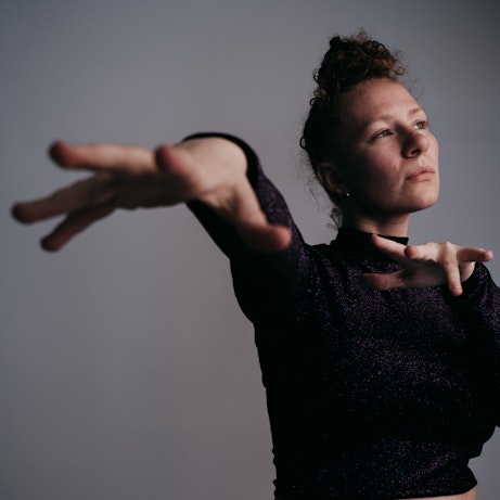 Jemma Freese stood in front of a grey background looking off to the right. Her hands and fingers are outstretched towards the camera.