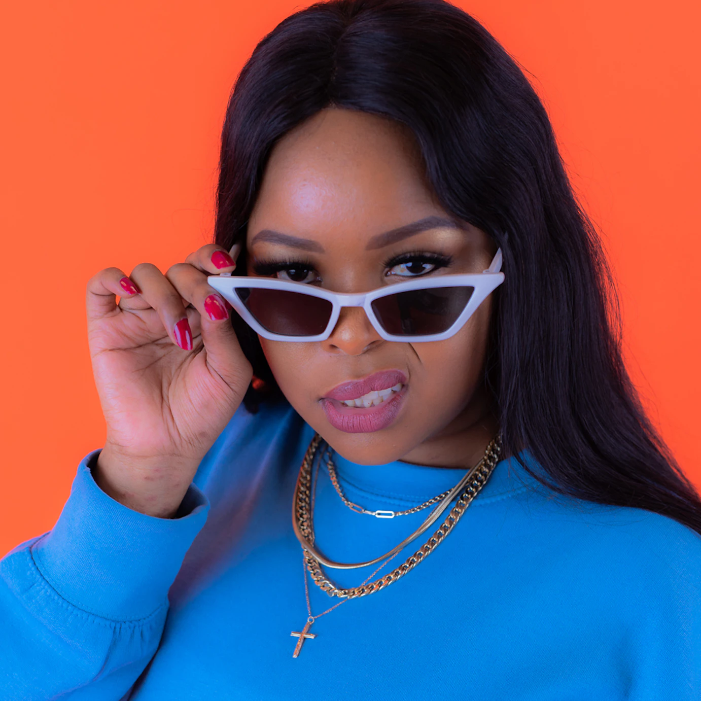 Ms. Dee looks directly into the camera with her shades lowered. She is wearing a bright blue jumper and gold chains, and is standing against a bright orange background.