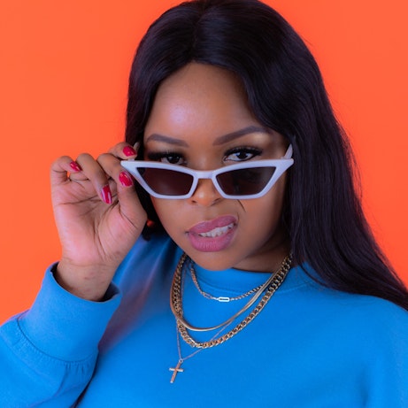 Ms. Dee looks directly into the camera with her shades lowered. She is wearing a bright blue jumper and gold chains, and is standing against a bright orange background. 