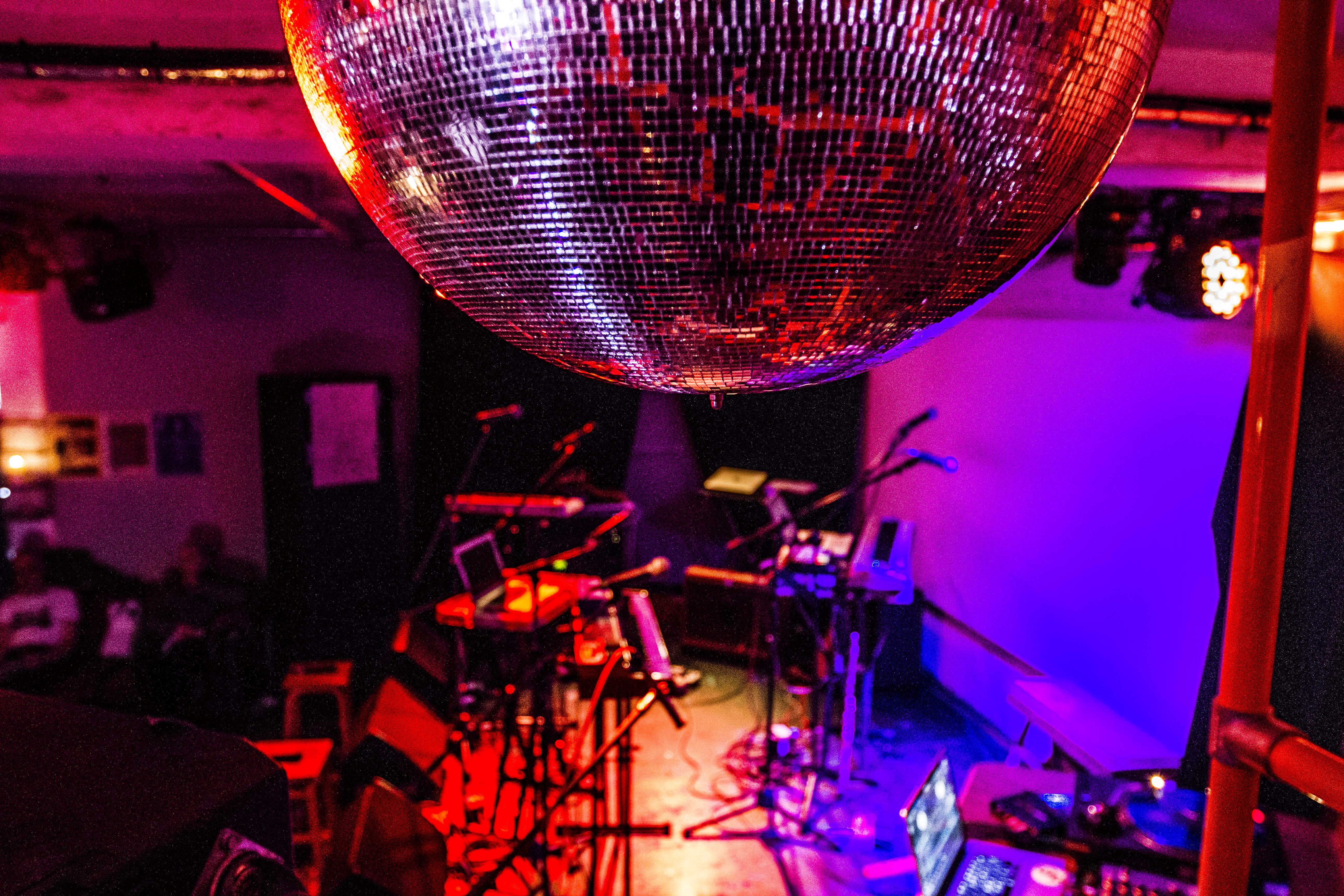 An empty stage cast in red, pink and purple light. Various instrument, laptops and stands are se up ready to be played. In focus just in front is a mirror ball on the ceiling.