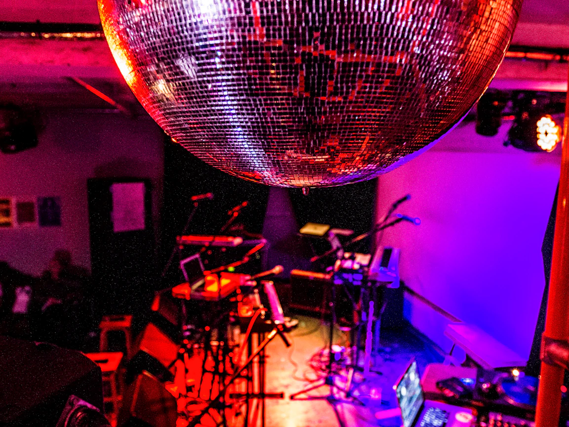 An empty stage cast in red, pink and purple light. Various instrument, laptops and stands are se up ready to be played. In focus just in front is a mirror ball on the ceiling.