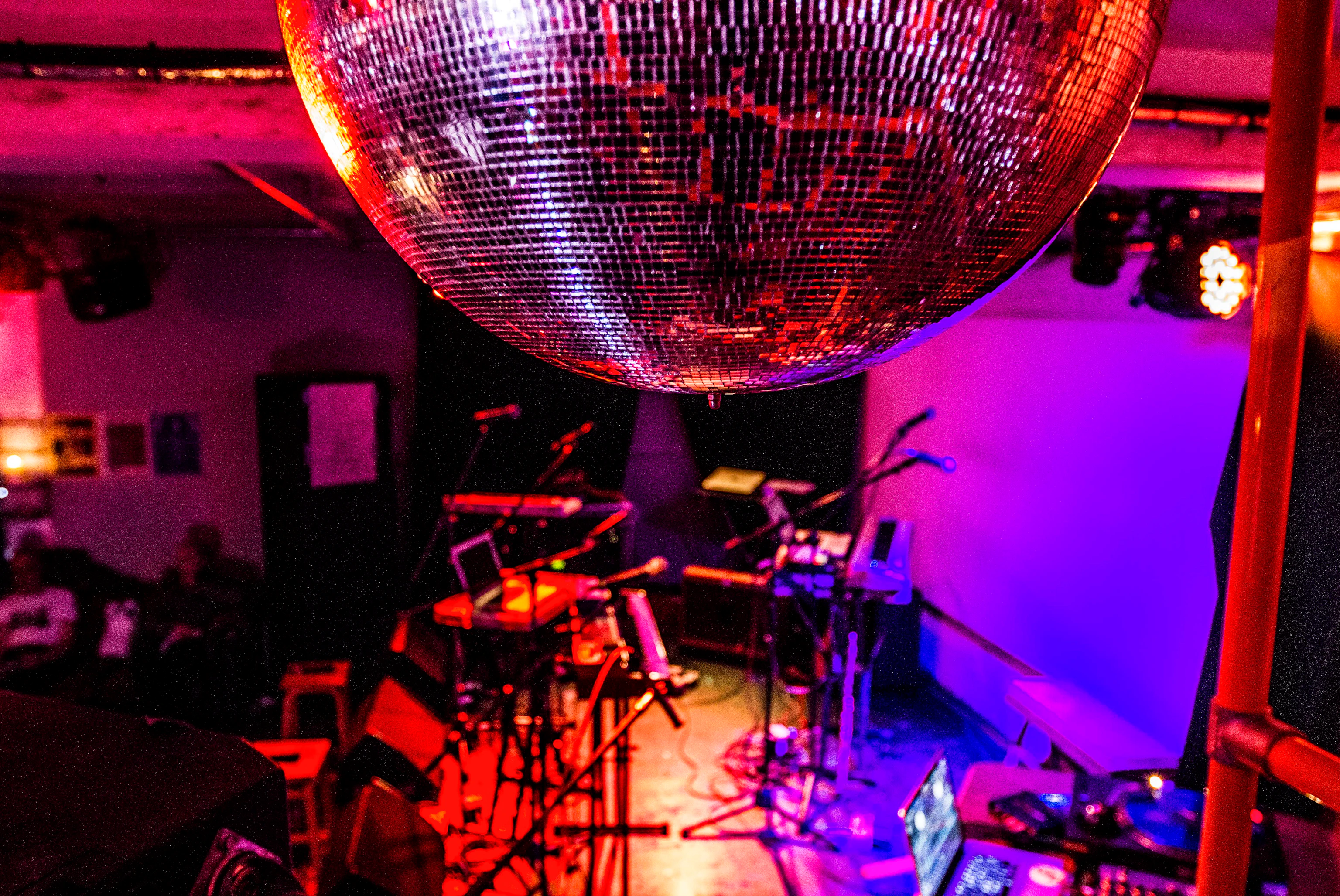 An empty stage cast in red, pink and purple light. Various instrument, laptops and stands are se up ready to be played. In focus just in front is a mirror ball on the ceiling.