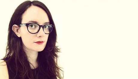 Laura Snapes stands to the left on an eggshell white background looking into the camera. She is wearing glasses and her dark hair flows down either side of her torso.