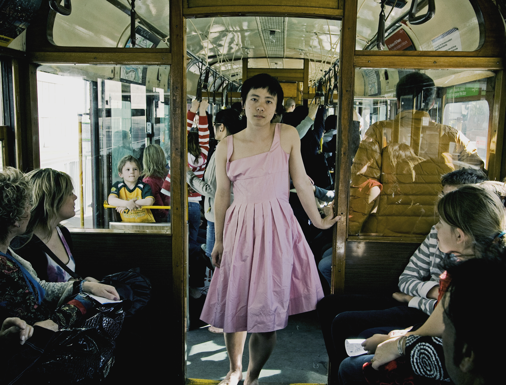 Jian Yi stood in the doorway of a busy bus looking into the camera. They are leant against the doorframe and are dressed in a light pink dress.