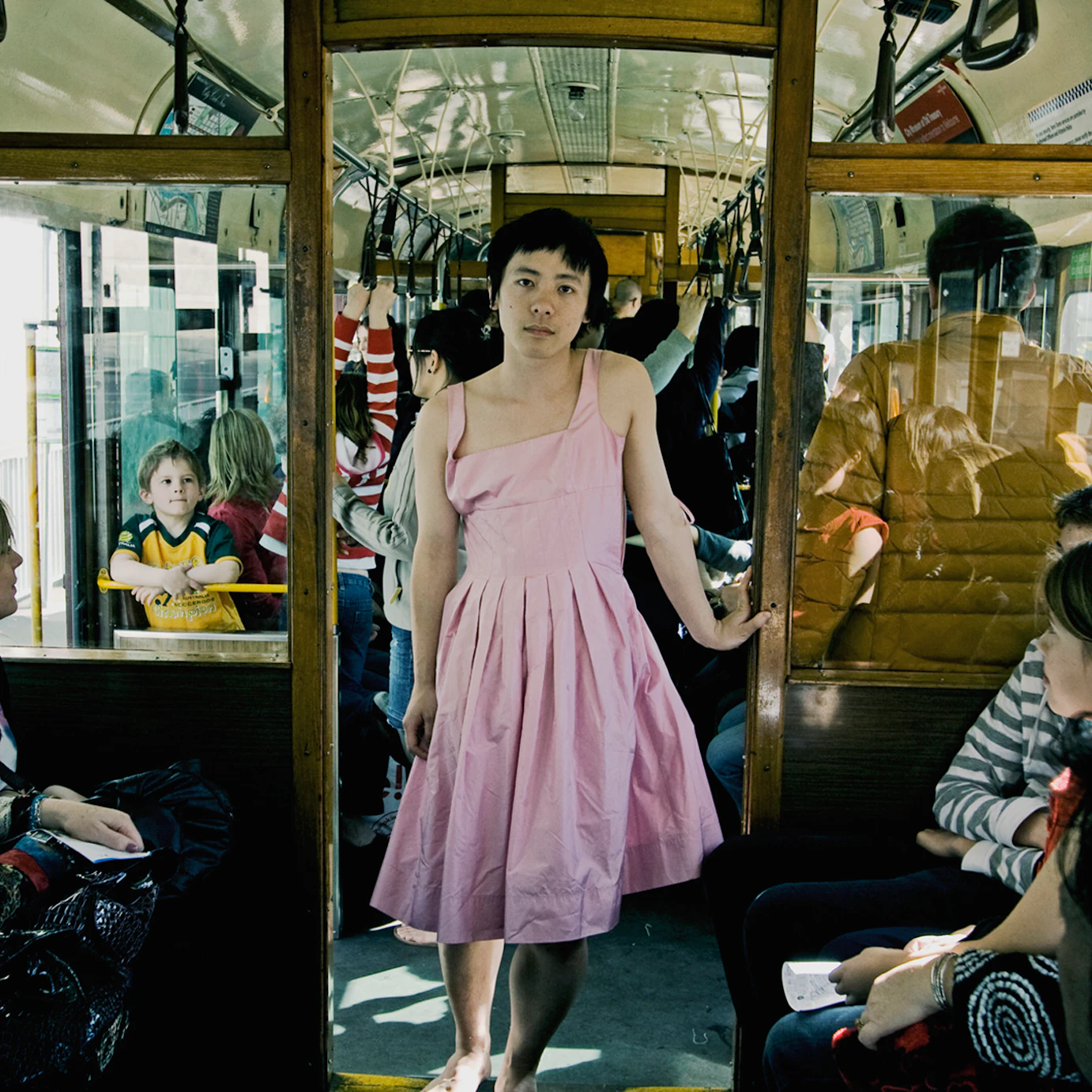 Jian Yi stood in the doorway of a busy bus looking into the camera. They are leant against the doorframe and are dressed in a light pink dress.