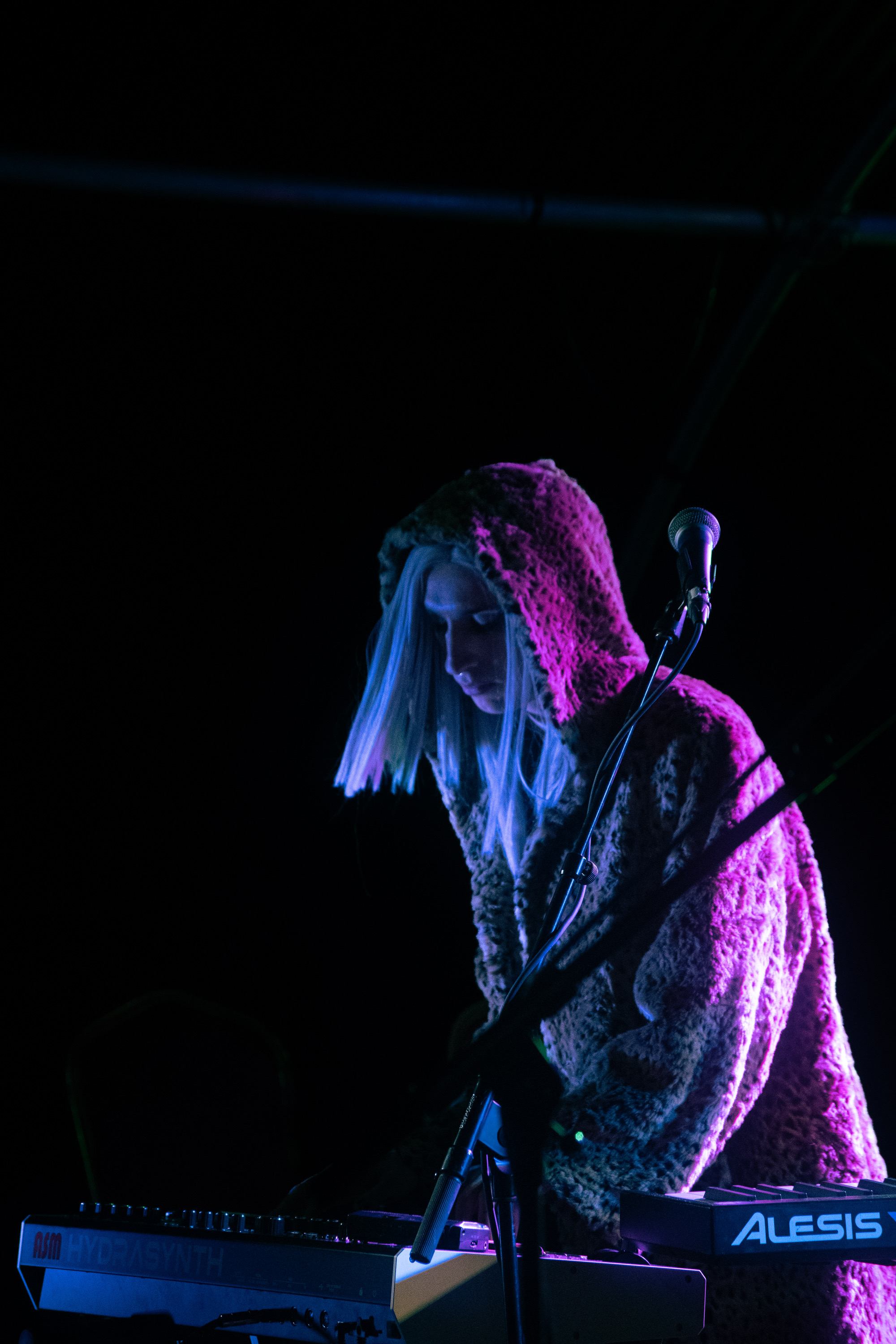 Phoenix Rousiamanis performing in a dark space. They are wearing a fluffy coat with the hood up and pink/purple light shines behind them.