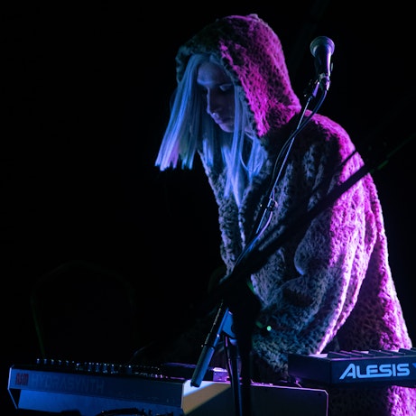 Phoenix Rousiamanis performing in a dark space. They are wearing a fluffy coat with the hood up and pink/purple light shines behind them.