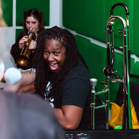 Hannabiell Sanders laughing in a music studio. Fee behind her is playing a trumpet.