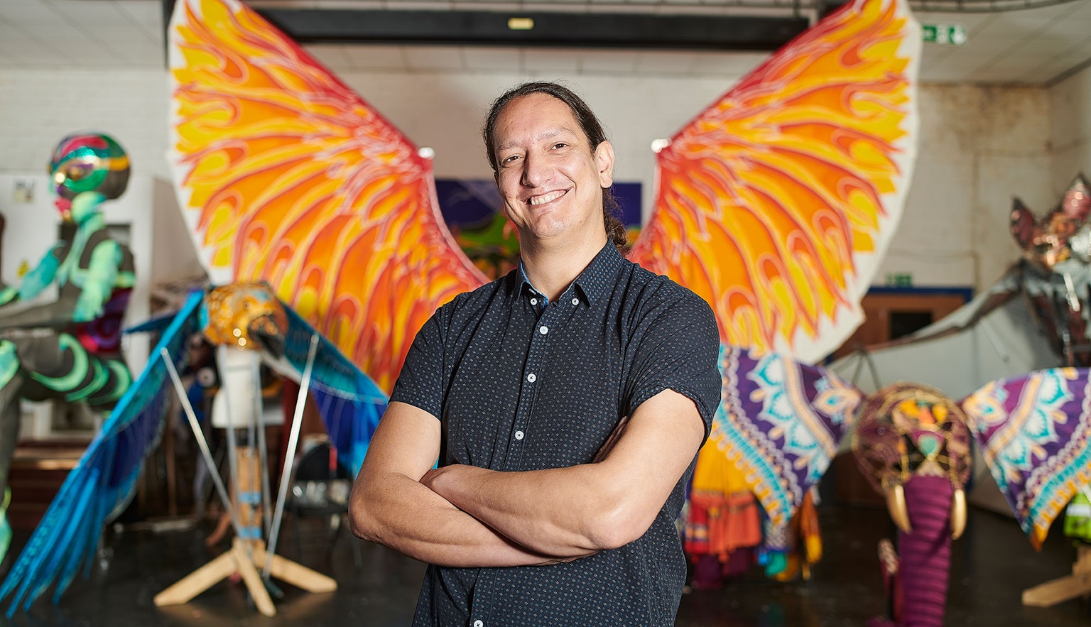 Leon Patel with his arms folded looking into the camera and smiling. Behind him are various multicoloured parade puppets in the shape of different animals.