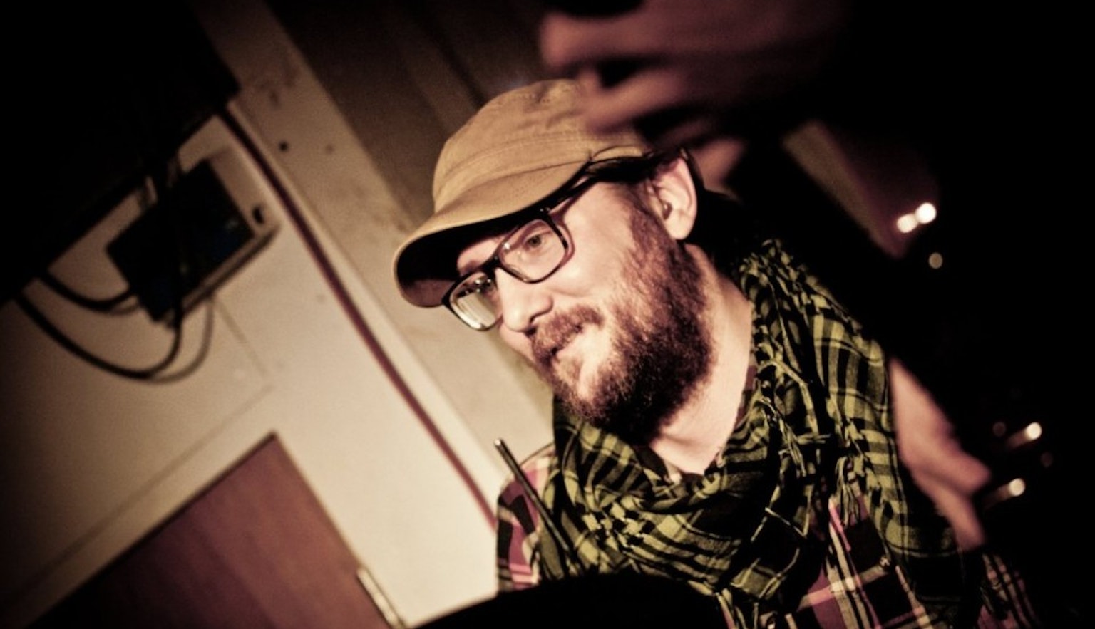 A close up of Phill Howley's face in a studio. You can just about see in the corners of the image that he's behind a drumset. He is wearing a beige cap, black glasses and a checkered scarf and shirt.