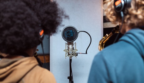 Two saxophonists with headphones on standing in a studio and playing into a microphone.