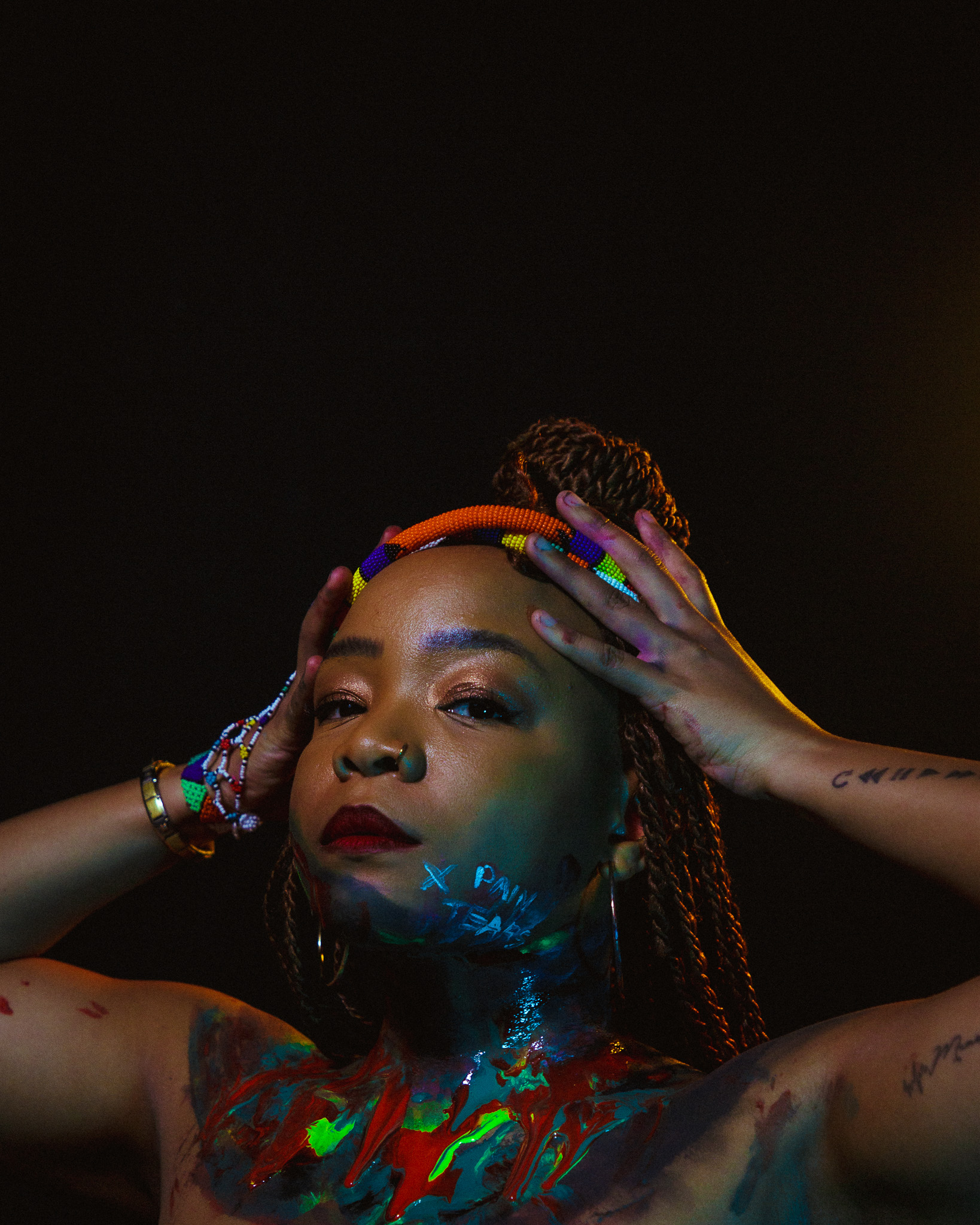 Artist Lebo is covered in various colours of paint across her torso and chin. Her hands are on her head and she's looking into the camera.