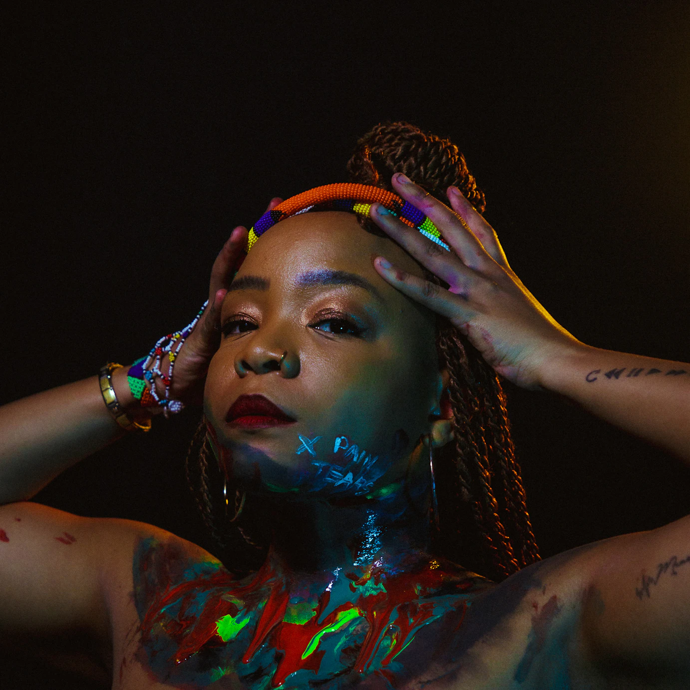Artist Lebo is covered in various colours of paint across her torso and chin. Her hands are on her head and she's looking into the camera.