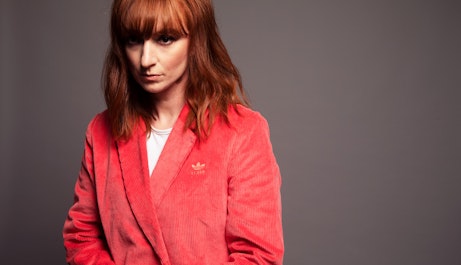 LoneLady stood looking into the forward distance wearing a bright red Adidas blazer on a grey backdrop.