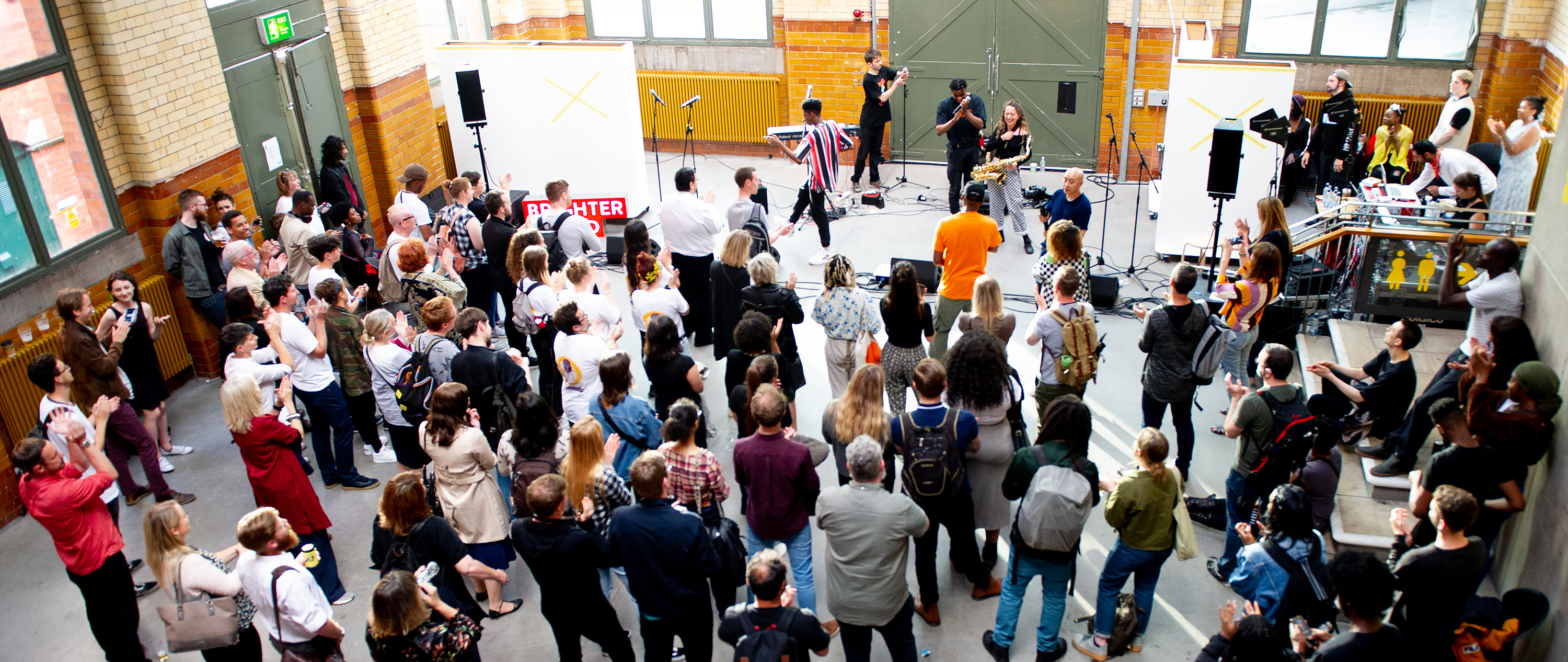 A crowd shot of the DISRUPT residency performance. There is no stage. Just microphones and other music equipment set up around the performers. The crowd have gathered in front of them, as a buskers crowd in the street might.