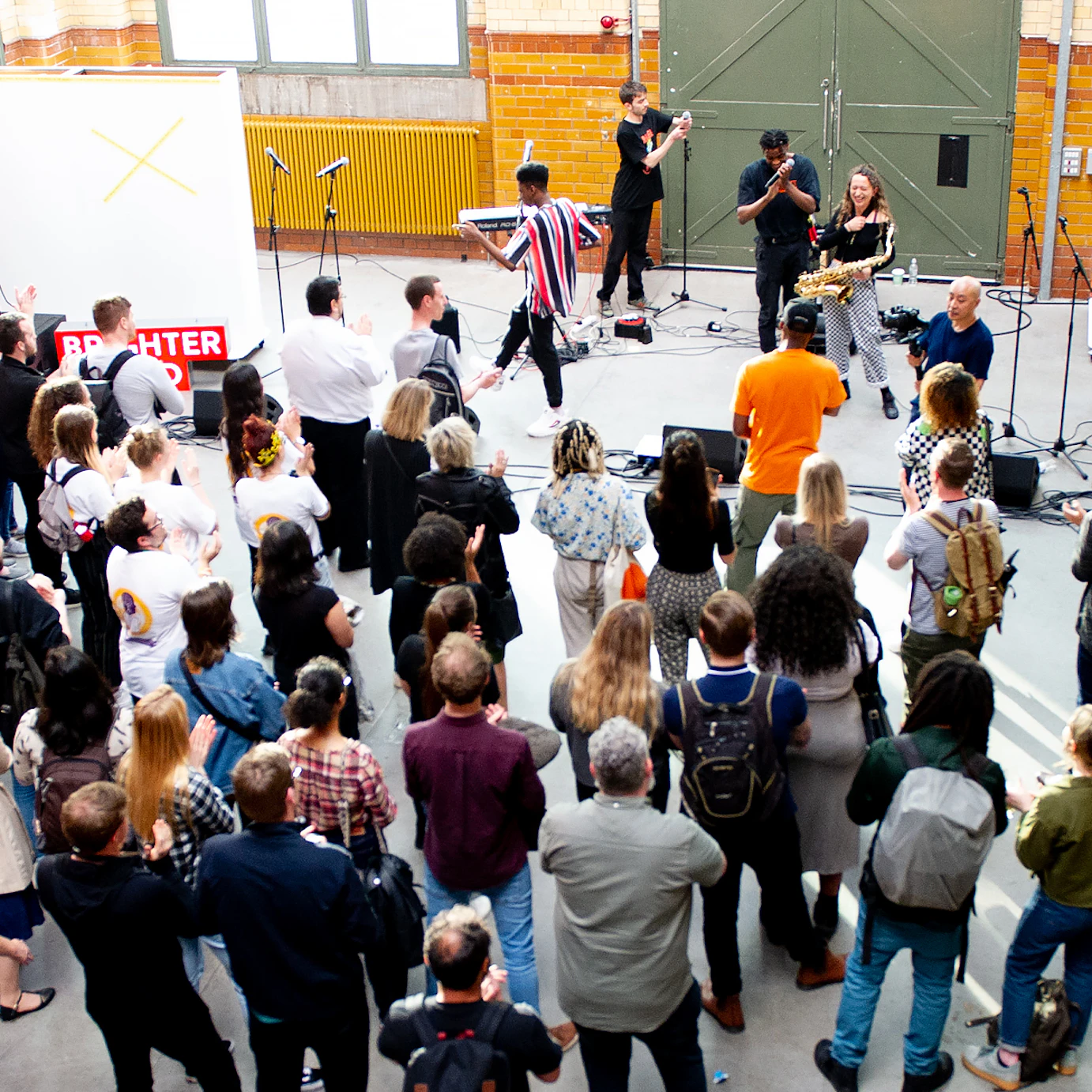 A crowd shot of the DISRUPT residency performance. There is no stage. Just microphones and other music equipment set up around the performers. The crowd have gathered in front of them, as a buskers crowd in the street might.