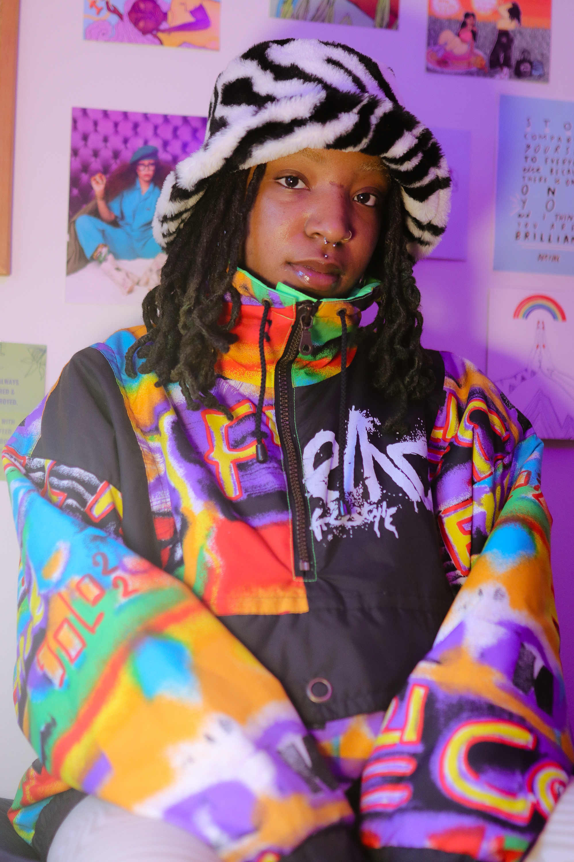 TU3SDAY sat by a wall with various colourful cartoons and images stuck on it. They're wearing a fluffy zebra patterned bucket hat and a baggy coat zipped up to their chin with multicoloured graffiti printed all over.