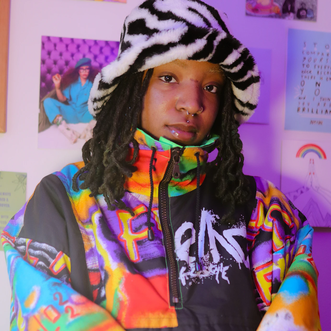 TU3SDAY sat by a wall with various colourful cartoons and images stuck on it. They're wearing a fluffy zebra patterned bucket hat and a baggy coat zipped up to their chin with multicoloured graffiti printed all over.