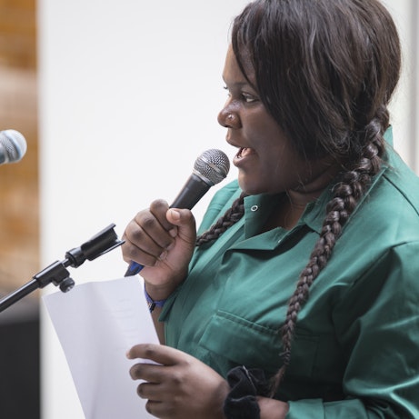 A residency artist dressed in a dark green boiler suit holding a sheet of paper and speaking into a microphone.