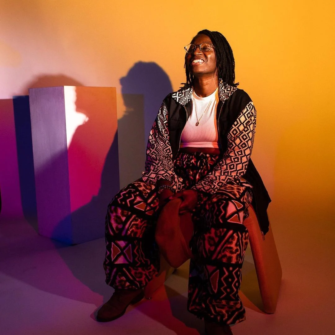 NikNak smiling and sat in a white studio space, lit by pink, orange and yellow lighting.
