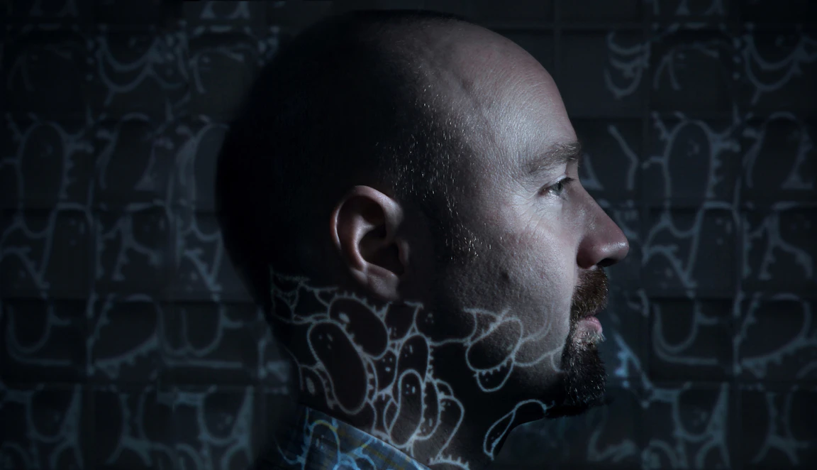 Dimly lit side profile of Mr Scruff with a projection of his illustrations which cover the background and his neck.