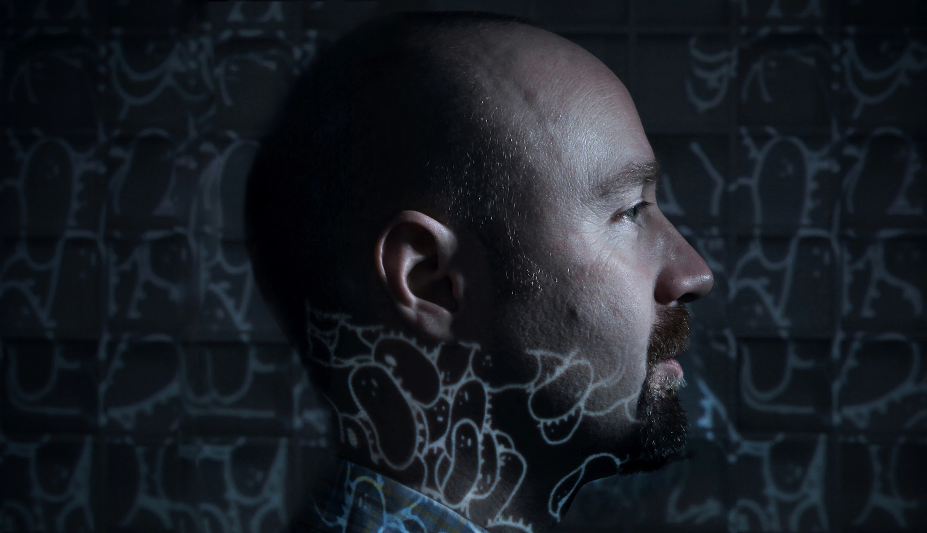 Dimly lit side profile of Mr Scruff with a projection of his illustrations which cover the background and his neck.