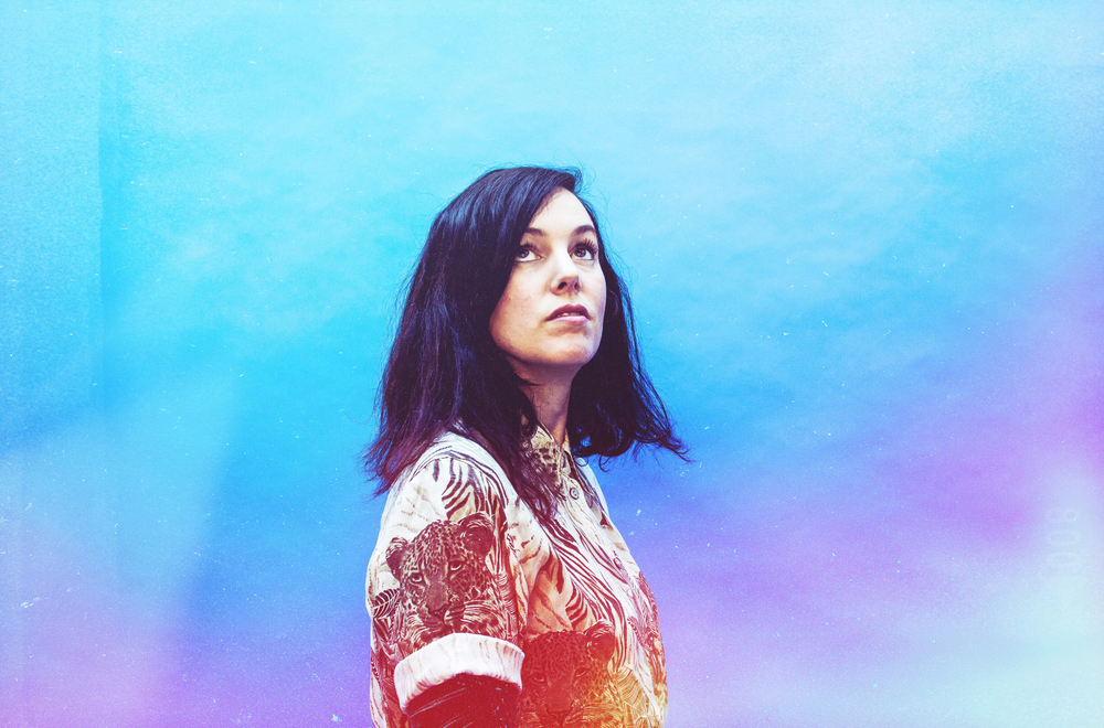 Anna Meredith stood in the centre looking up towards something unseen. The background is a mixture of blues and purples that seep behind and in front of Anna, transitioning to warm reds, oranges and yellows on her torso.