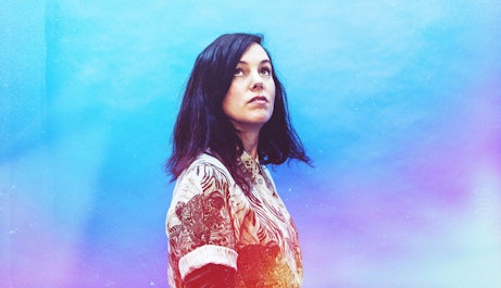 Anna Meredith stood in the centre looking up towards something unseen. The background is a mixture of blues and purples that seep behind and in front of Anna, transitioning to warm reds, oranges and yellows on her torso.