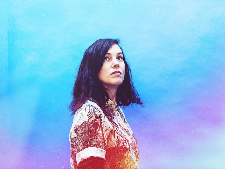 Anna Meredith stood in the centre looking up towards something unseen. The background is a mixture of blues and purples that seep behind and in front of Anna, transitioning to warm reds, oranges and yellows on her torso.