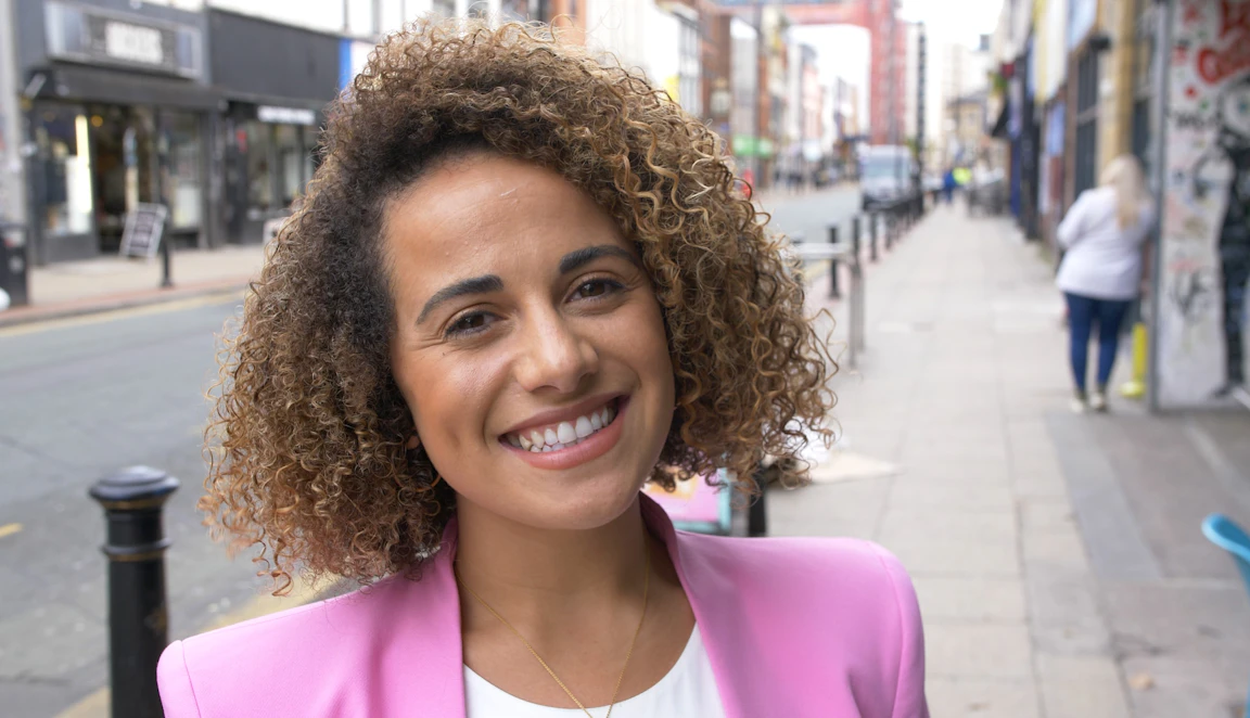 Chesqua stands on a Manchester street. She is looking straight into the camera with a beaming smile.