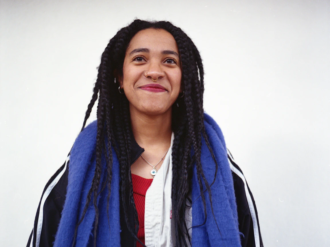 Kinaya smiles at the camera and wears an Adidas jacket and purple scarf. Kinaya is standing in front of a white background.