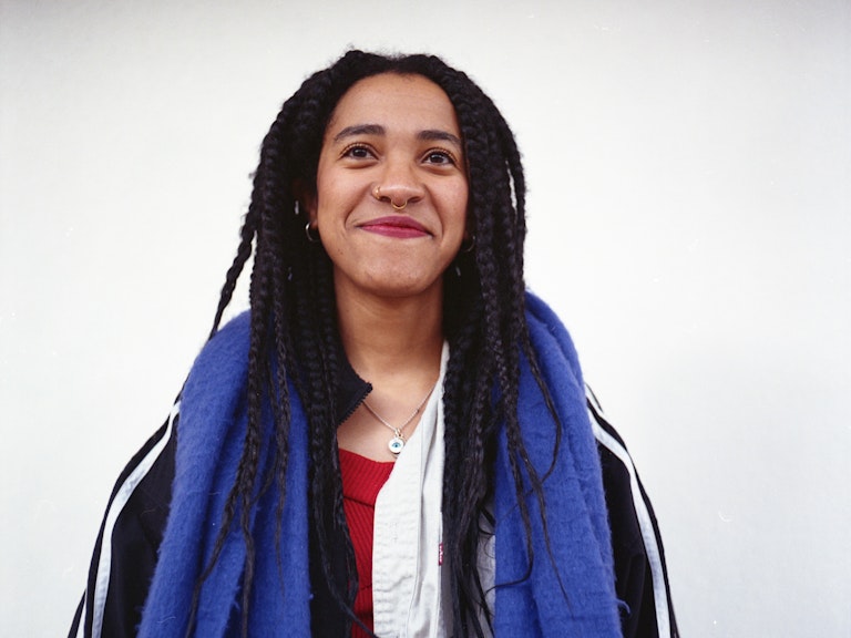 Kinaya smiles at the camera and wears an Adidas jacket and purple scarf. Kinaya is standing in front of a white background. 