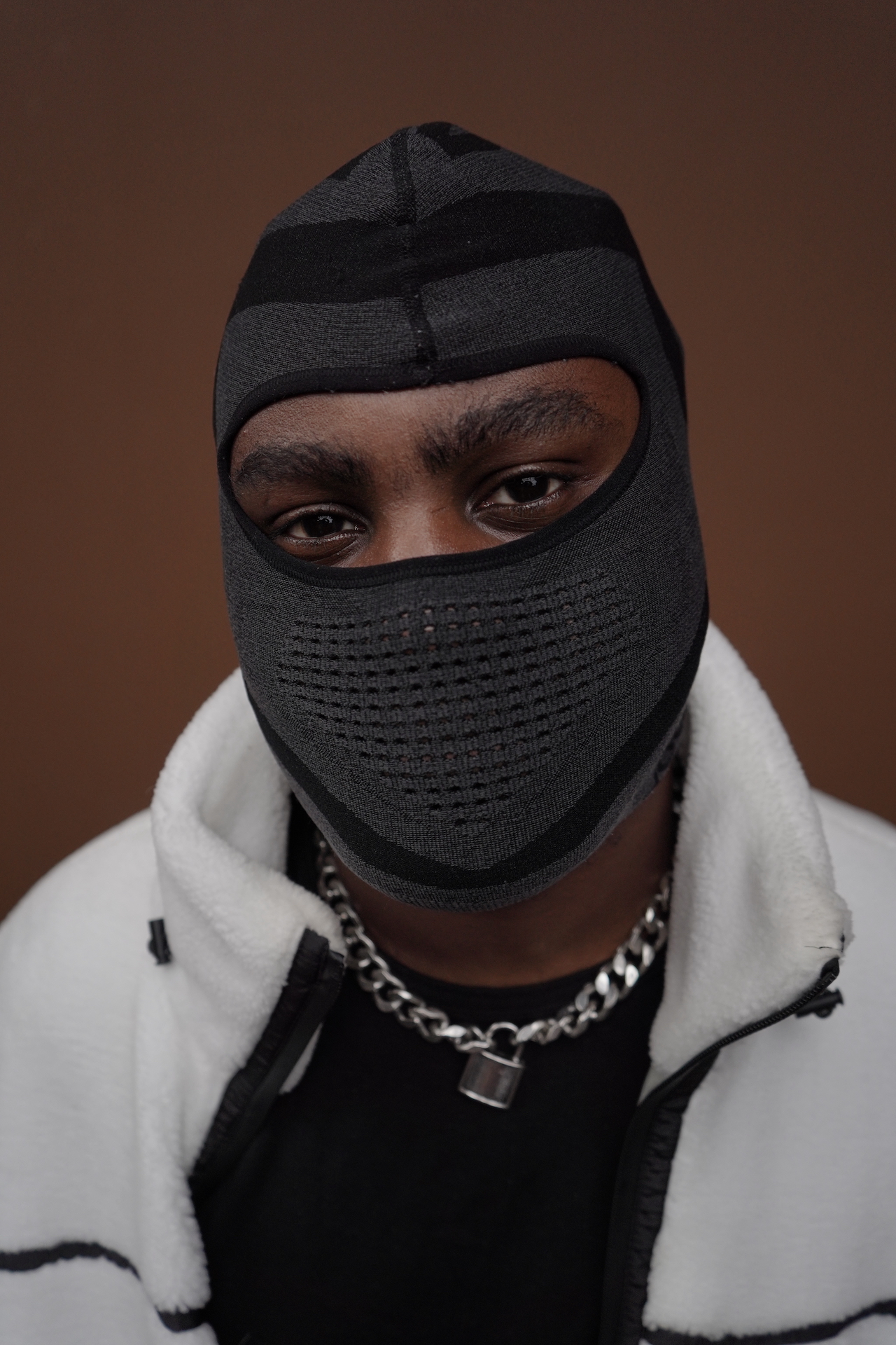VRSYJNES on a brown background looking into the camera. His face is hidden by a grey and black balaclava. He's wearing a white and black fleece with a silver chain around his neck.