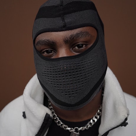 VRSYJNES on a brown background looking into the camera. His face is hidden by a grey and black balaclava. He's wearing a white and black fleece with a silver chain around his neck.
