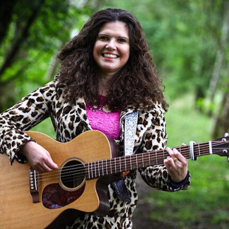 Elena Piras stood in woodland playing a guitar and smiling into the camera.