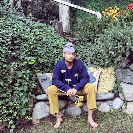 Khaleb Brooks sat in a garden on a bench made of large stones piled on top of one another. He's wearing a beanie, blue jumper with daisies, yellow trousers and no shoes. He's holding two sunflowers.