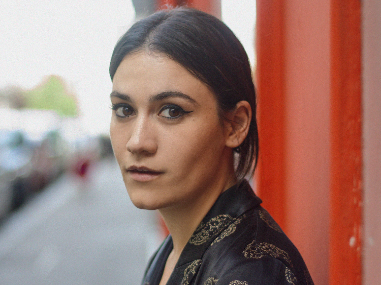 Nadine Shah is standing on the pavement against a red painted building and looks directly at the camera. Nadine is wearing a black blazer with gold embroidery.