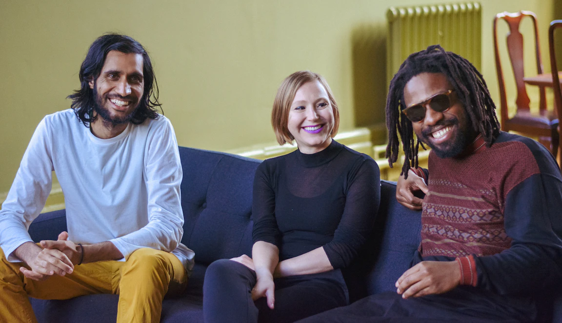 Balraj, Kamila and Tunde sit together on a sofa. They are all smiling.