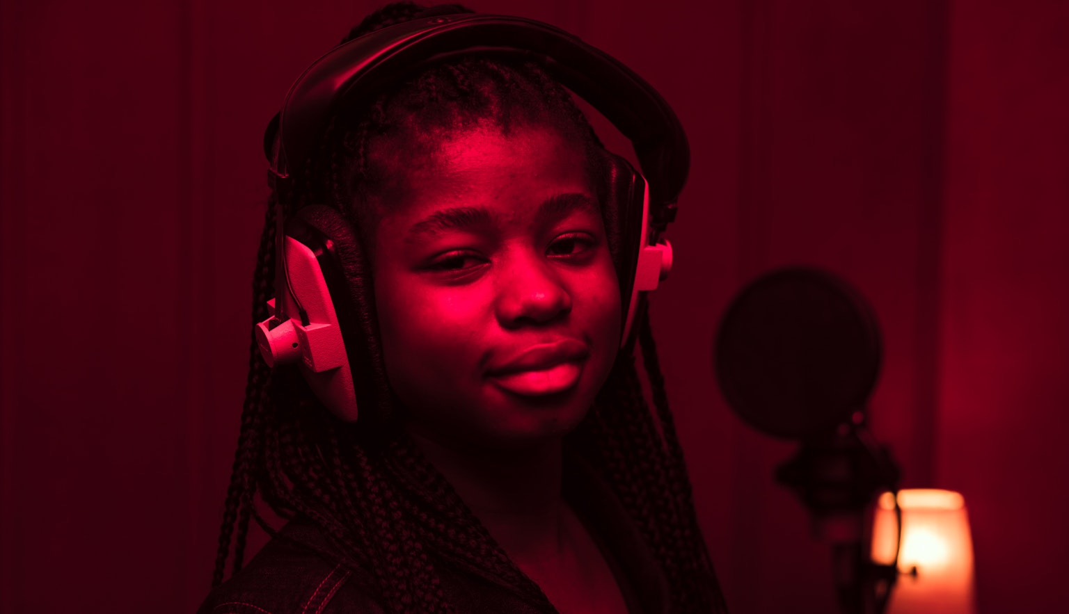 A young black girl in a recording studio with headphones on. The room has a deep red glow to it.