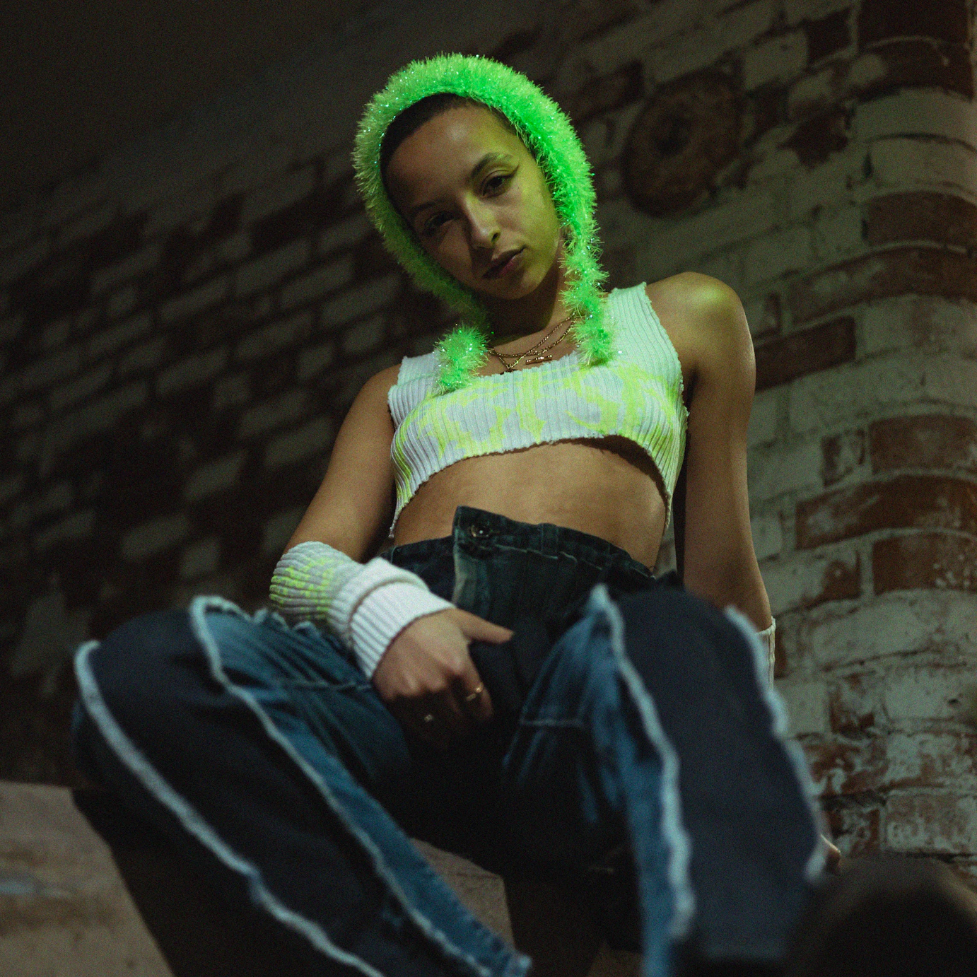 Karis Jade sat on a ledge with a brick structure behind her looking down into the camera.