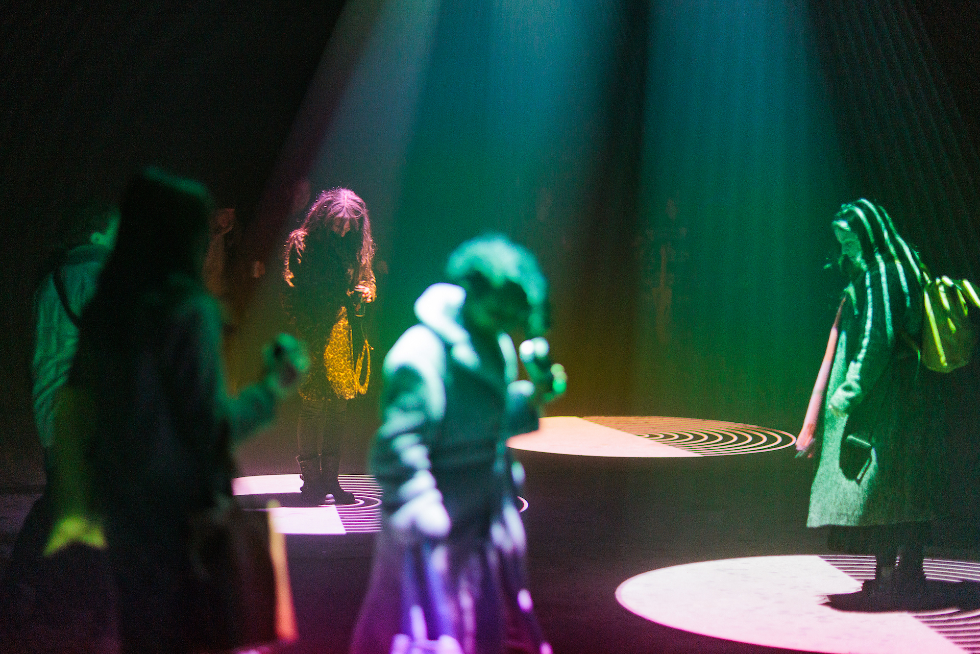 In a dark space, various people are illuminated in rainbow colours from projections shining down from above. On the floor they project rows of circles rippling out and growing in size.