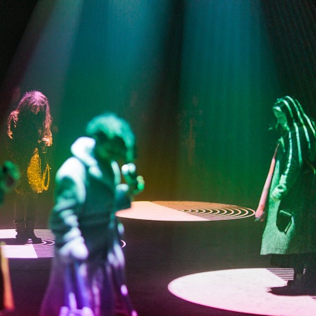 In a dark space, various people are illuminated in rainbow colours from projections shining down from above. On the floor they project rows of circles rippling out and growing in size.