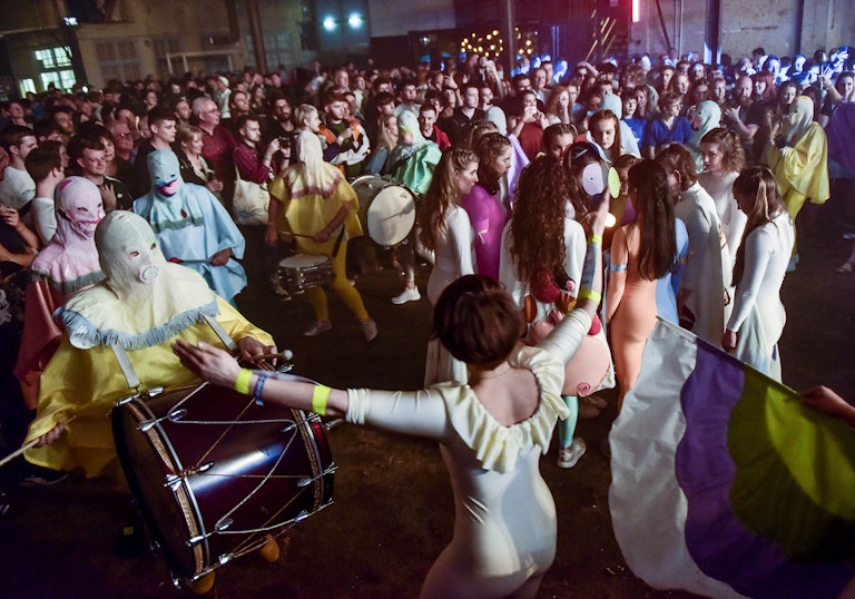 A large crowd are watching a performance in an open space with drummers, dancers and a flag.