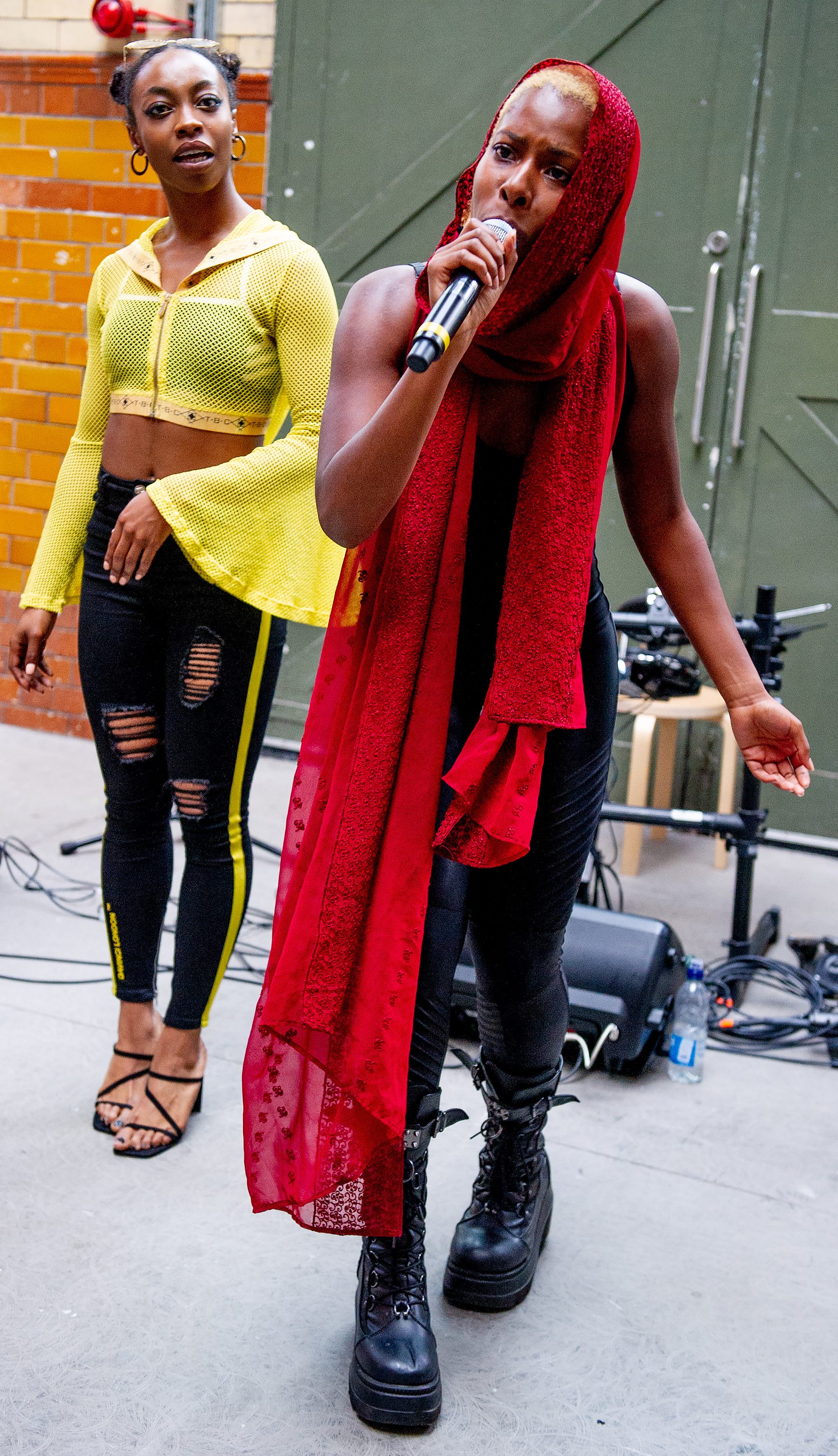 Olivia May speaking into a microphone with a red scarf wrapped around her neck and head. Sarah Angel stands behind her wearing yellow and black.