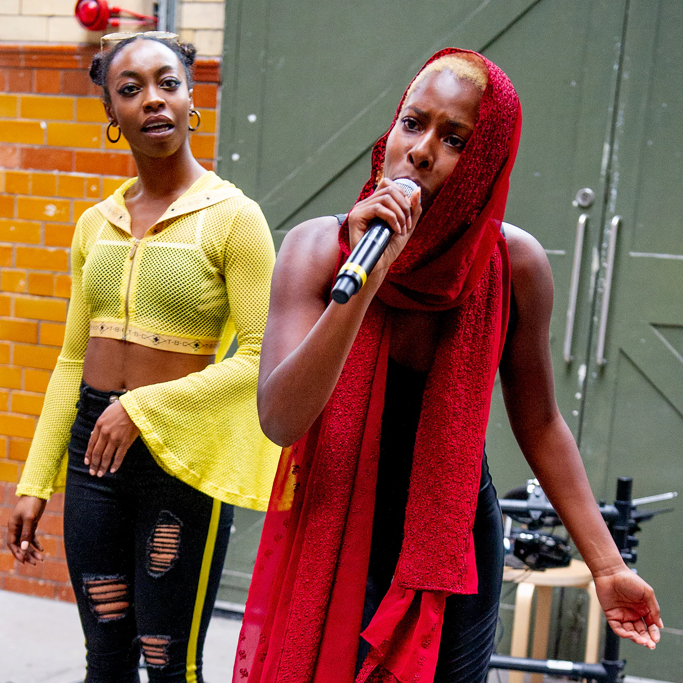 Olivia May speaking into a microphone with a red scarf wrapped around her neck and head. Sarah Angel stands behind her wearing yellow and black.