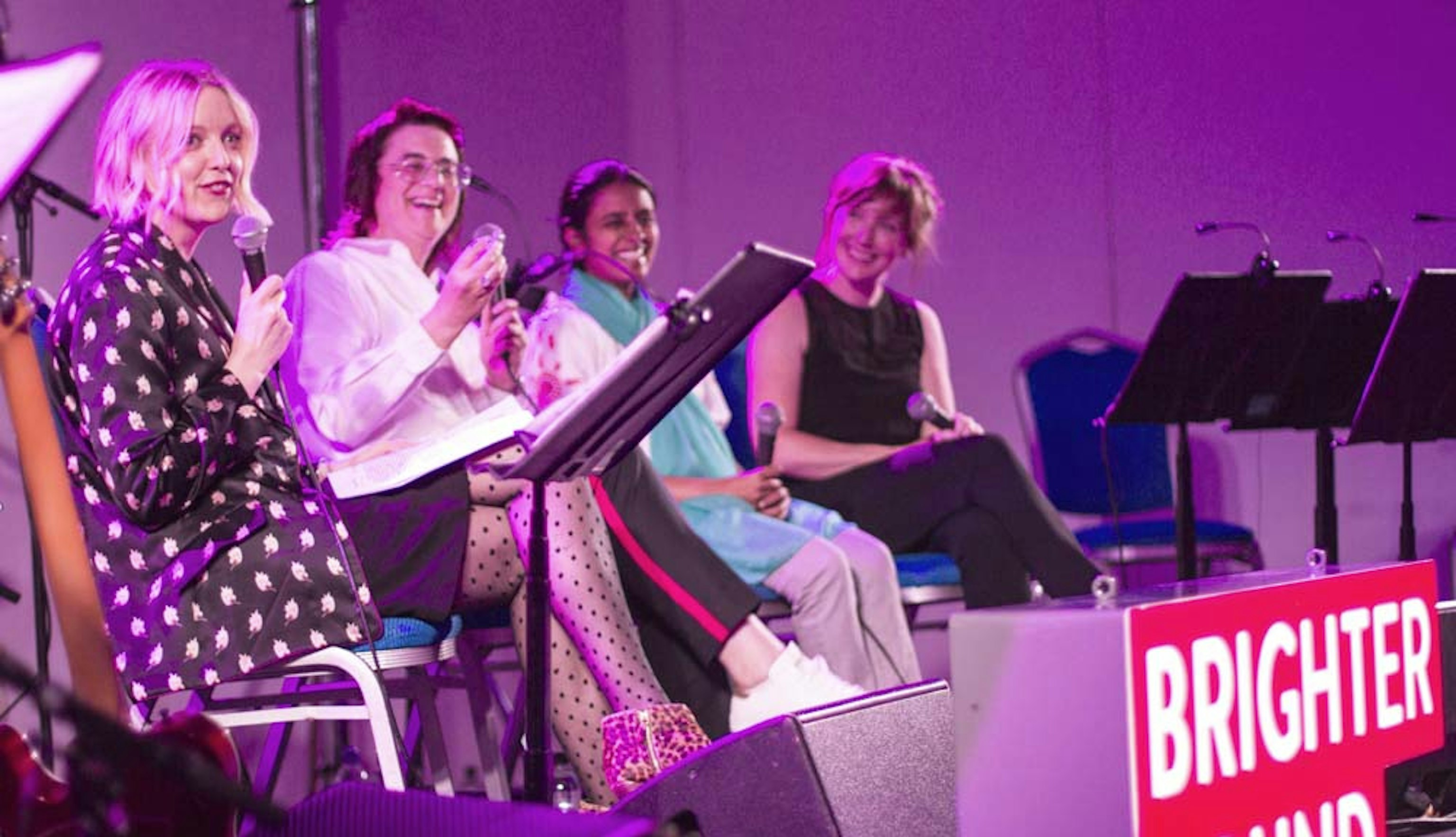 Panel from Adventures in Flatland. They are all sitting down, smiling and holding microphones. There is a glow from the purple light. In front of them is a Brighter Sound lightbox in the shape of our old logo.