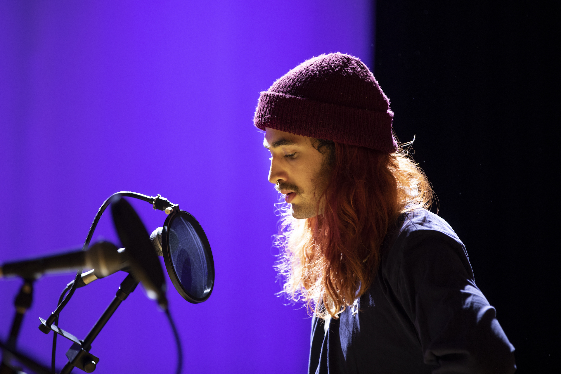 A participant wearing a beanie over their long red hair looking down whilst facing a microphone.