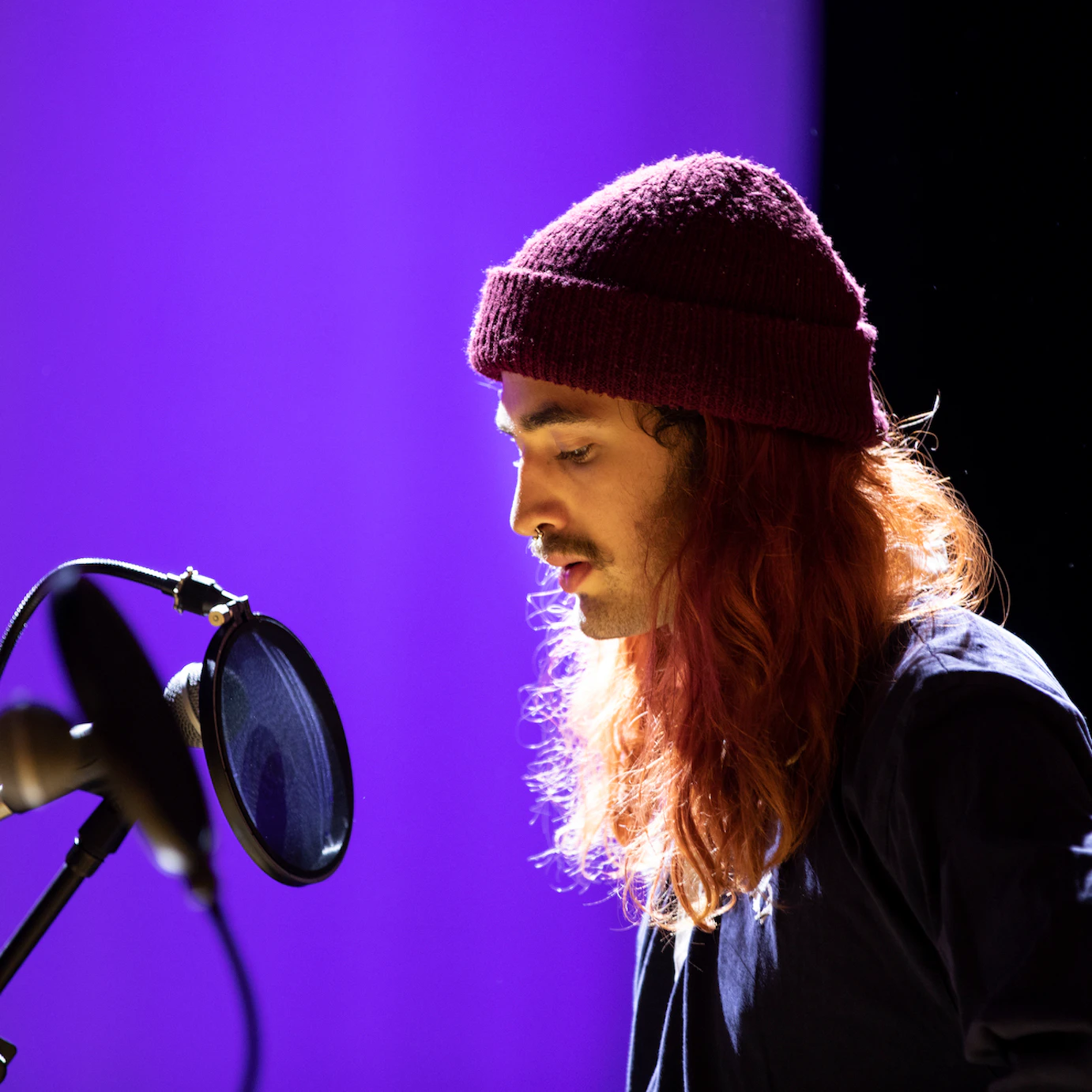 A participant wearing a beanie over their long red hair looking down whilst facing a microphone.