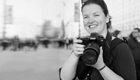 Rachel Bywater stands to the right smiling into the camera. She is holding a camera with both hands just below her face. A background of passersby is out of focus.